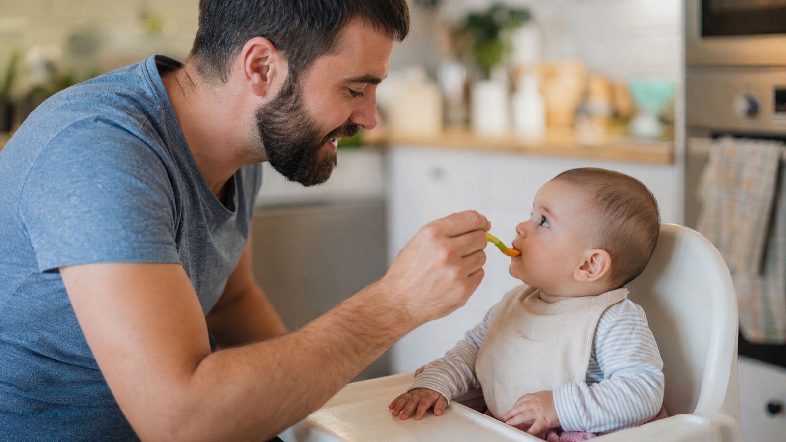 Vader in mosterdgele trui voert zijn baby een lepel groentepuree in de kinderstoel